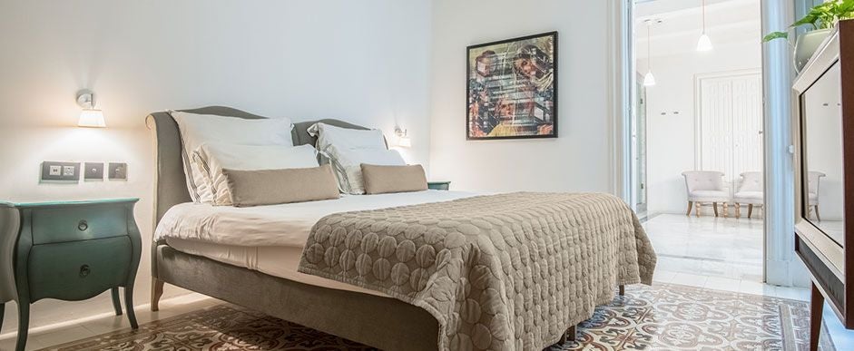 Elegant boutique hotel suite with soft neutral tones, plush king bed, designer furniture, and ornate Maltese architectural details in warm evening light