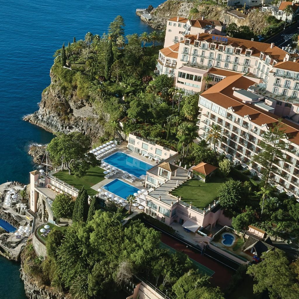 Pink-hued luxury hotel with ornate balconies and terraces cascading down a dramatic oceanside cliff in Madeira, surrounded by lush gardens