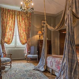 Elegant French château hotel room with antique furnishings, soft cream walls, classic four-poster bed, and large windows overlooking manicured park grounds