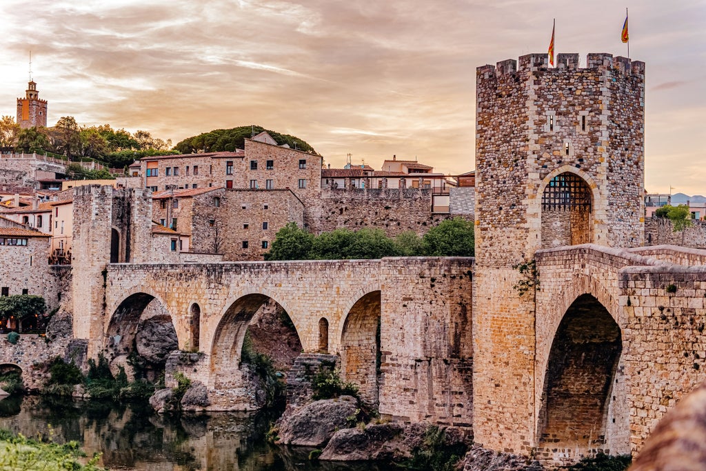 Stone medieval bridge spanning a river, leading to fortified town with ancient stone buildings and towers in Besalú, Catalonia, Spain at golden hour