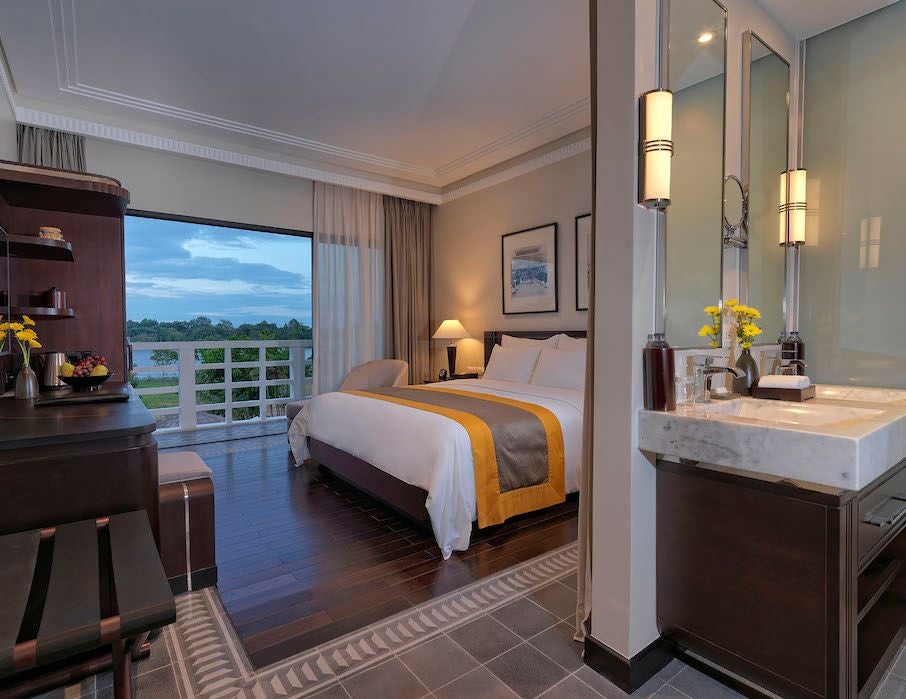 Elegant Vietnamese riverside hotel room with classic colonial design, wooden furnishings, and expansive windows overlooking serene Perfume River in Hue