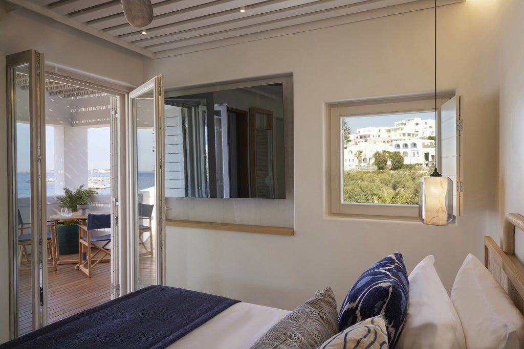 Modern seaside hotel room with plush double bed, white linens, and floor-to-ceiling windows offering panoramic ocean or side views in Greece