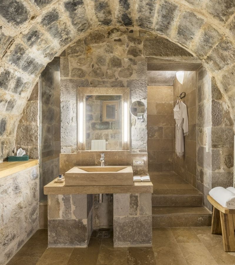 Elegant stone-walled suite with plush white bedding, arched windows, and traditional Turkish decor overlooking Cappadocia's unique volcanic landscape