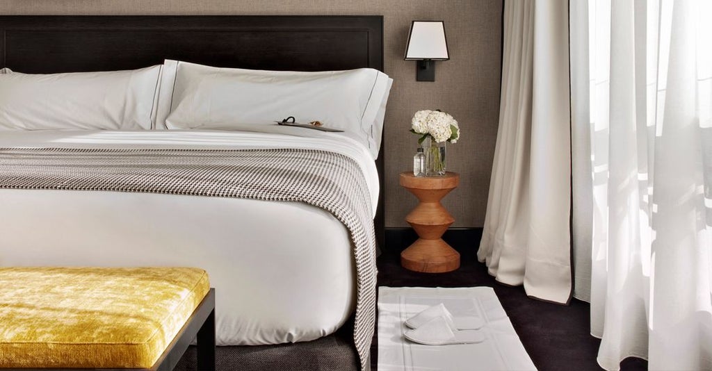 Luxurious premium hotel room with modern design, plush king-sized bed, elegant neutral tones, and large windows overlooking Madrid's cityscape at sunset