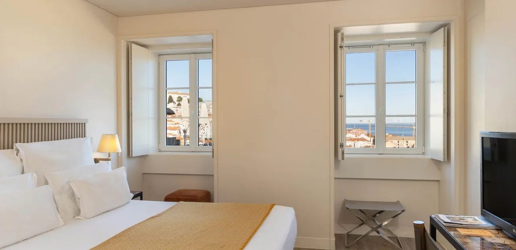 Elegant minimalist hotel room in Lisbon's Alfama district, featuring white walls, wooden floor, crisp white linens, and large window with city view