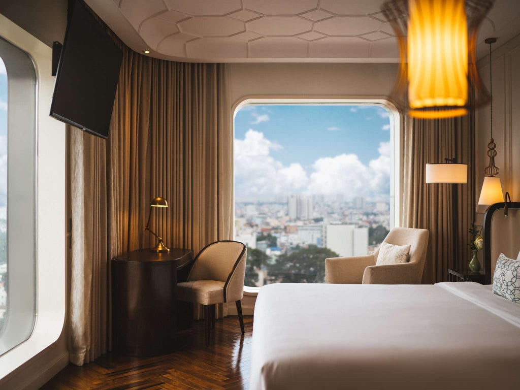 Modern corner hotel room with floor-to-ceiling windows offering city views, plush king bed, elegant furnishings and soft neutral decor