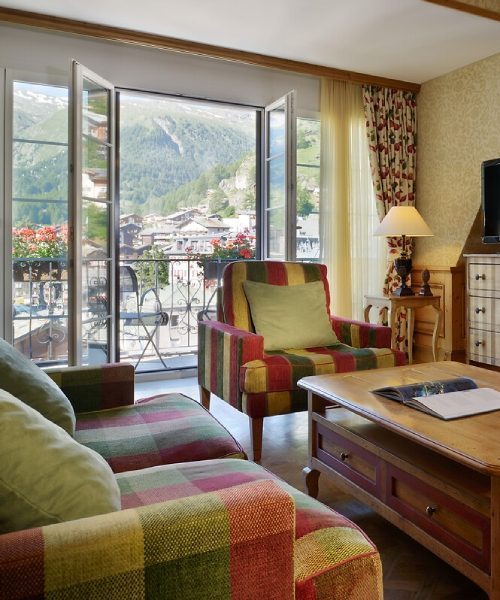 Elegant Swiss alpine suite with panoramic mountain views, featuring dark wood beams, plush furnishings, and a private terrace