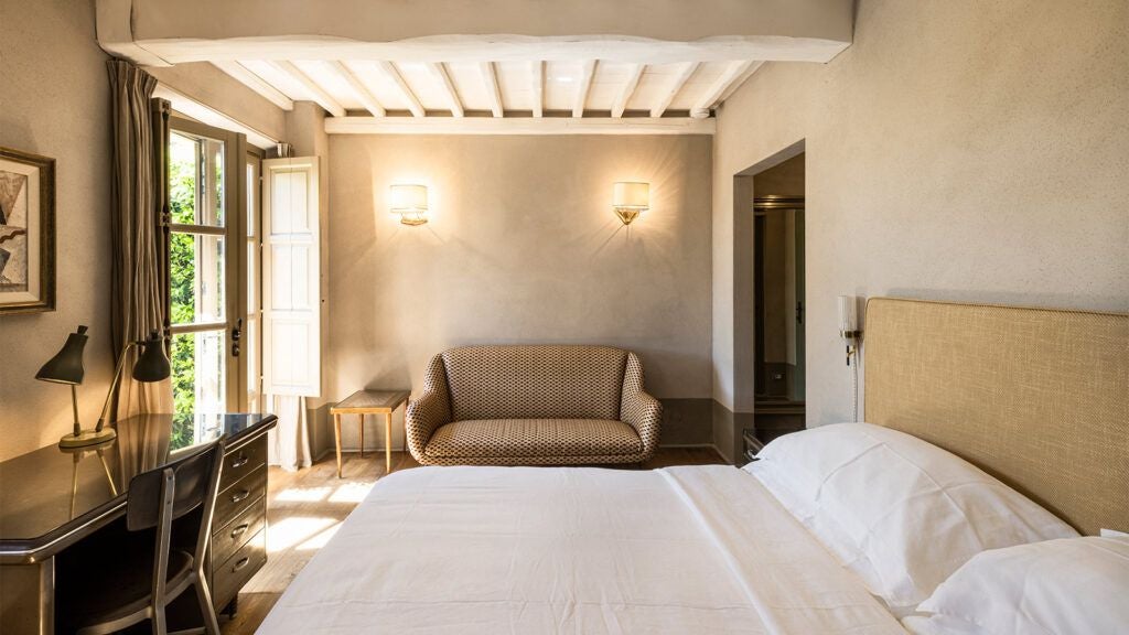 Elegant Italian hotel room with soft neutral tones, plush white bedding, minimalist wooden furniture, and panoramic countryside view through large windows