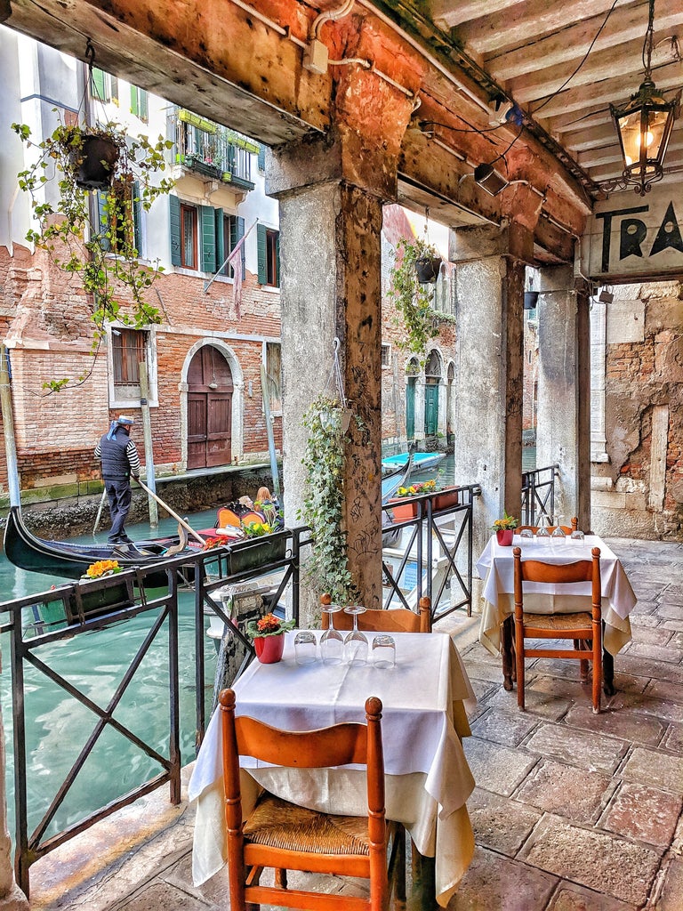 Traditional wooden gondola gliding through narrow Venice canal, elegant couple enjoying champagne beside ornate buildings with floral balconies