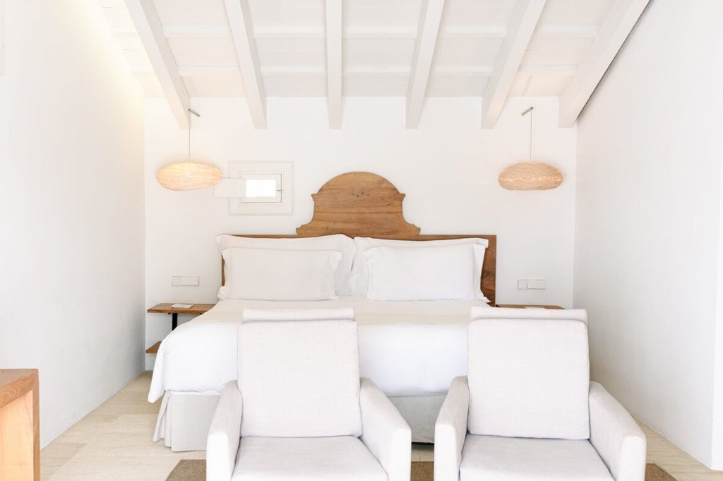 Elegant whitewashed stone hotel room with minimalist design, wooden bed, crisp linens, and panoramic window overlooking Mediterranean countryside in Menorca, Spain