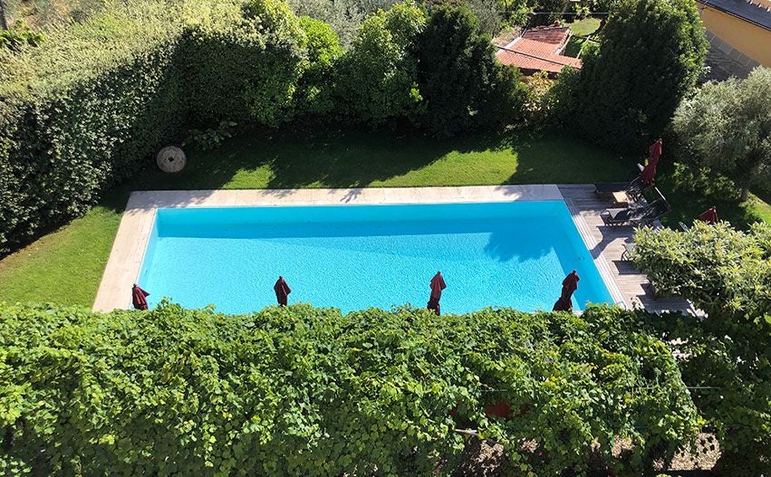 Elegant Italian villa bedroom with French doors opening to a sparkling blue pool, lush garden, and traditional terracotta tiled rooftops in background