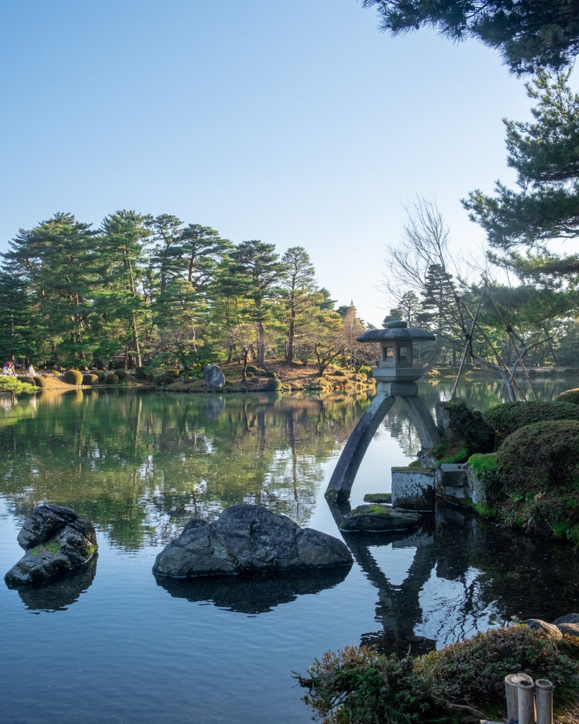 Traditional wooden bridge over steaming hot spring leads to luxurious Japanese ryokan surrounded by lush autumn maples in Kaga Onsen