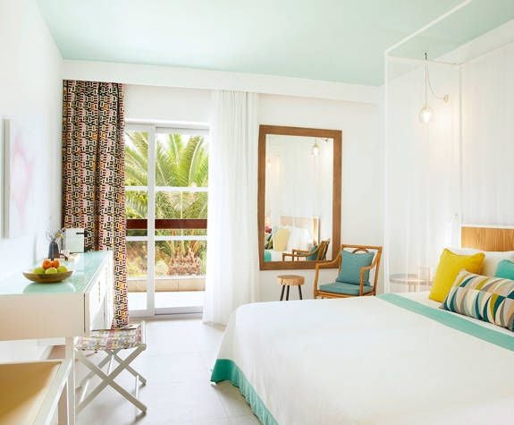 Elegant seaside hotel room at Eagles Palace with plush white bed, blue-teal Aegean Sea view, modern minimalist decor, and sunlight streaming through large windows