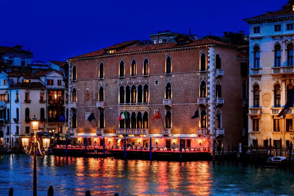 Ornate 15th-century Venetian palace hotel with marble columns and balconies overlooking the Grand Canal at sunset, gondolas passing by