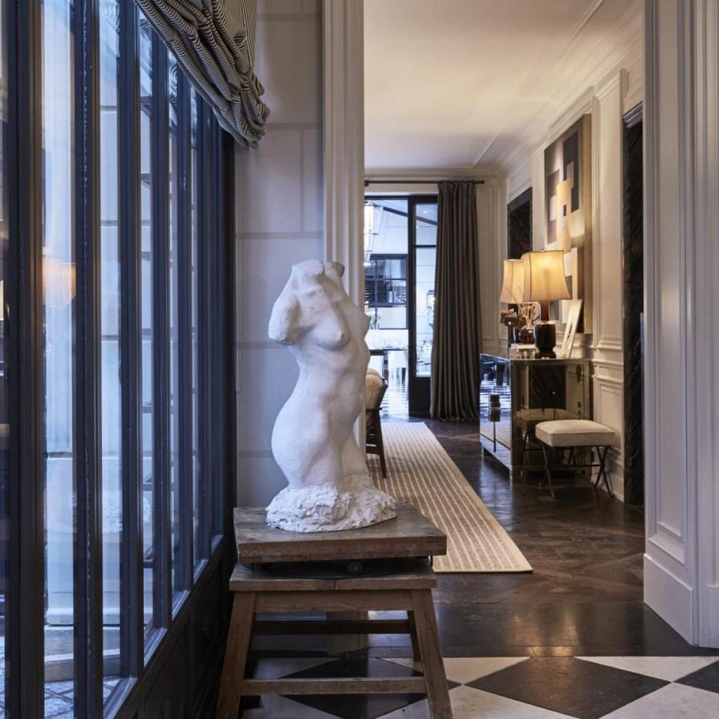 Elegant Parisian boutique hotel bedroom with white marble fireplace, plush cream-colored furniture, and soft natural lighting from large windows