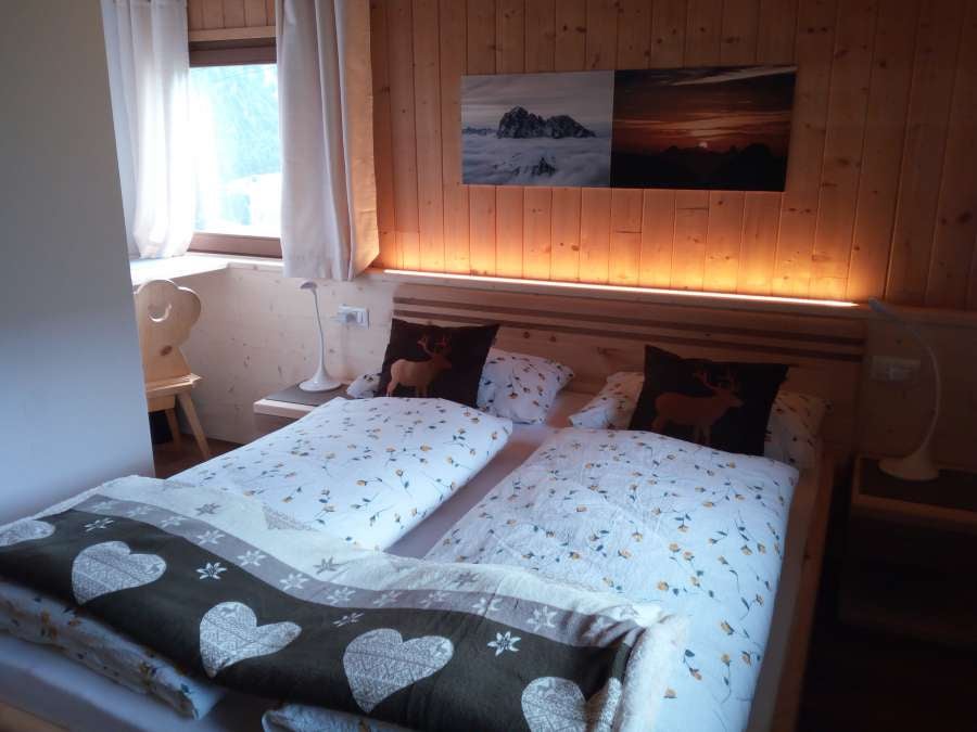 Cozy twin bedroom with rustic wooden walls, modern bedding, floor-to-ceiling windows framing dramatic Dolomite mountain views, soft lighting