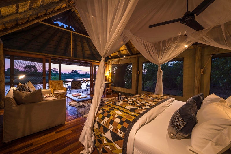 Elevated safari lodge at sunset with wooden deck overlooking African savanna, featuring plush outdoor lounge seating and ambient lighting