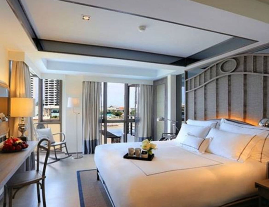 Modern hotel room with plush king bed, white linens, teak furnishings, floor-to-ceiling windows offering bright natural light, and Thai-inspired decor