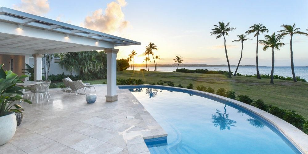 Luxurious Estate Suite at Jumby Bay, featuring white four-poster bed, wood floors, tropical decor, and panoramic ocean view through large windows