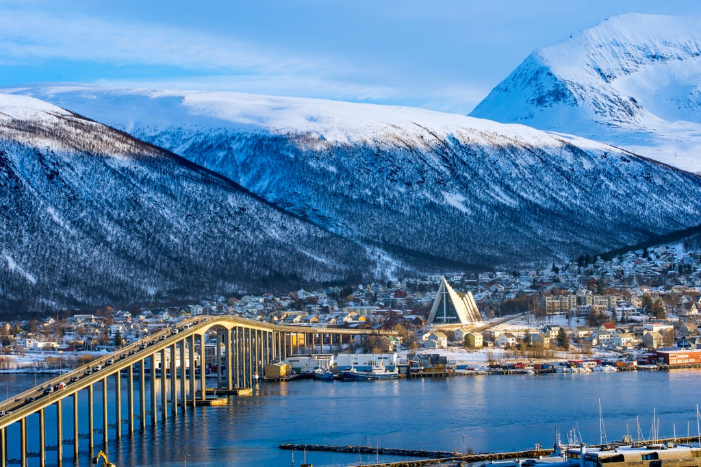 Modern wooden architecture of Arctic Cathedral illuminated at dusk, reflecting in fjord waters against snowy mountains in Tromsø