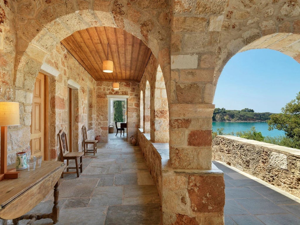 Sunlit stone villa bedroom with Mediterranean view, elegant wooden furnishings, and traditional Greek architectural details overlooking scenic landscape