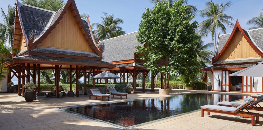 Spacious Thai villa bedroom with dark wood accents, white linens, floor-to-ceiling windows, and tropical garden views beyond terrace