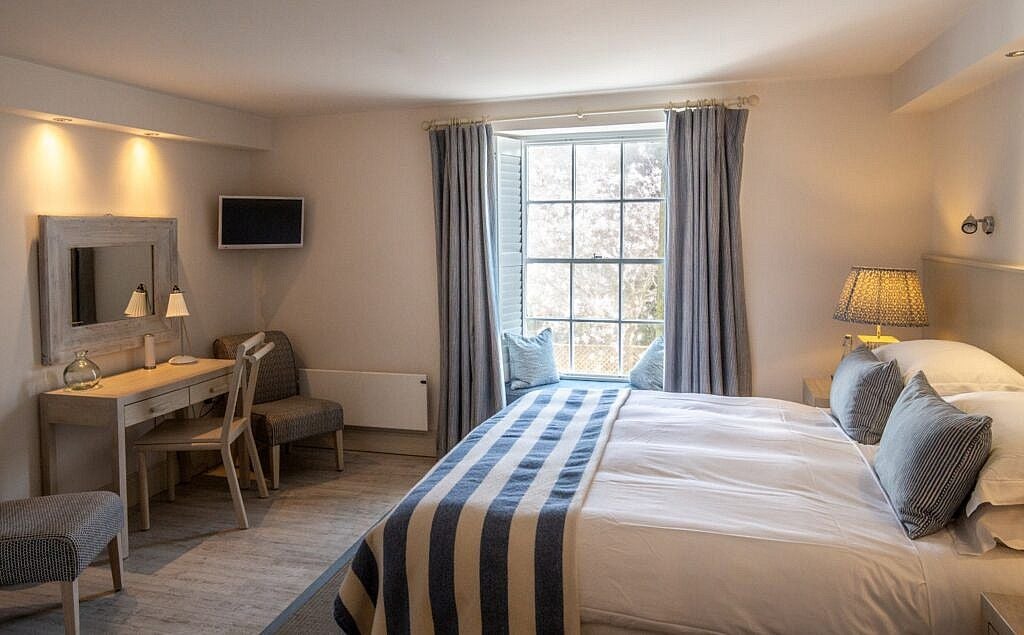 Cozy coastal village room at scenset hotel with crisp white bedding, nautical blue accents, and panoramic window overlooking charming seaside landscape