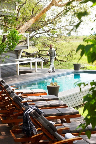 Elevated wooden walkway leads to luxurious tent-style safari lodges overlooking tranquil Okavango Delta at sunset, with palm trees