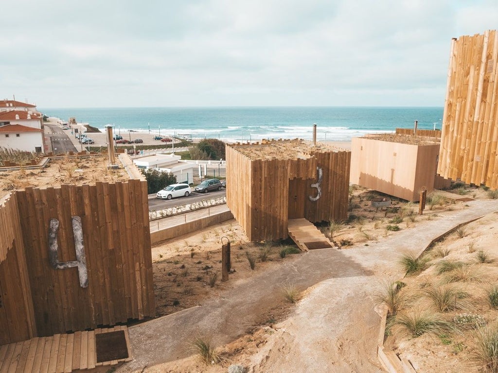 Modern minimalist bungalow with ocean view, wooden deck, white walls, and blue accents, overlooking rocky coastal landscape at Noah Surf House in Portugal.