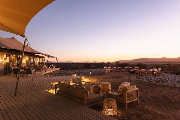 Luxury desert lodge with thatched-roof bungalows nestled against dramatic rust-colored Namibian landscape, featuring stark rocky terrain and distant mountain silhouettes