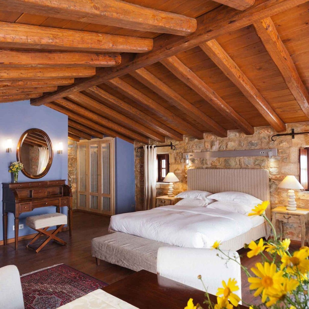 Elegant loft suite at coastal Greek hotel, featuring stone walls, wooden beams, plush white bedding, and panoramic view of azure Mediterranean Sea