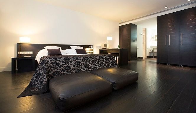 Elegant junior suite with plush white bedding, modern minimalist design, rich hardwood floors, and city view through expansive floor-to-ceiling windows in Valencia