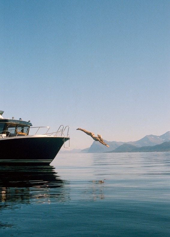 Elegant private boat cruising past towering cliffs in Norway's narrow fjord, sunlight illuminating turquoise waters and snow-capped peaks