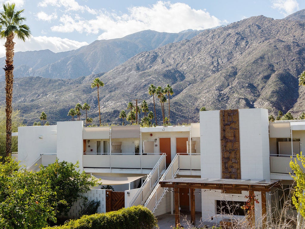 Modern retro-style hotel exterior with white geometric architecture, palm trees swaying against desert mountains at golden hour