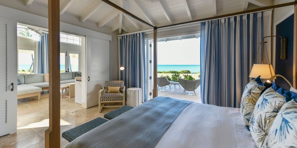 Luxurious Estate Suite at Jumby Bay, featuring crisp white linens, elegant wooden furnishings, and panoramic ocean views through floor-to-ceiling windows.