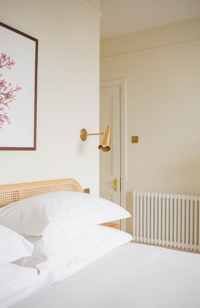 Modern luxury hotel room with elegant white bedding, soft neutral tones, minimalist wooden furniture, large window with natural light, and sophisticated coastal-inspired design elements.