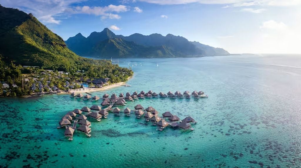 Over-water bungalows with thatched roofs at luxury resort in Moorea, stretching into turquoise lagoon surrounded by tropical palms