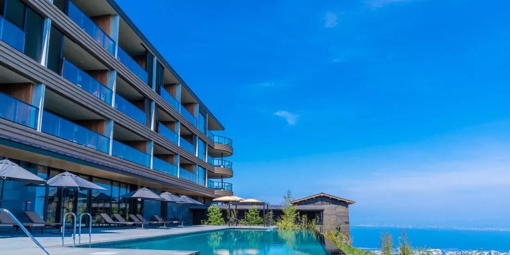 Modern luxury resort hotel with dramatic curved architecture, glowing at dusk against mountains overlooking Beppu's city lights