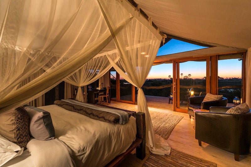 Elevated luxury safari tent with wooden deck overlooking vast Botswana wetlands, surrounded by lush palm trees at sunset