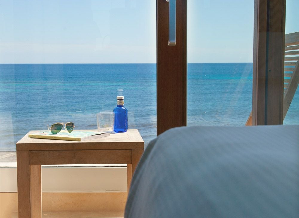 Luxurious beachfront hotel room with modern minimalist design, warm sunset colors, elegant coastal decor, and panoramic ocean view from private balcony