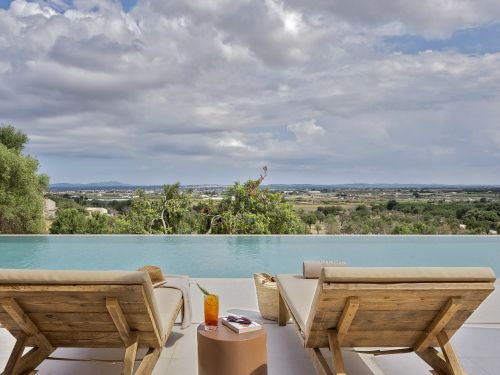 Luxurious Mediterranean-style villa with stone facade, lush green gardens, and infinity pool overlooking scenic Mallorca landscape at sunset