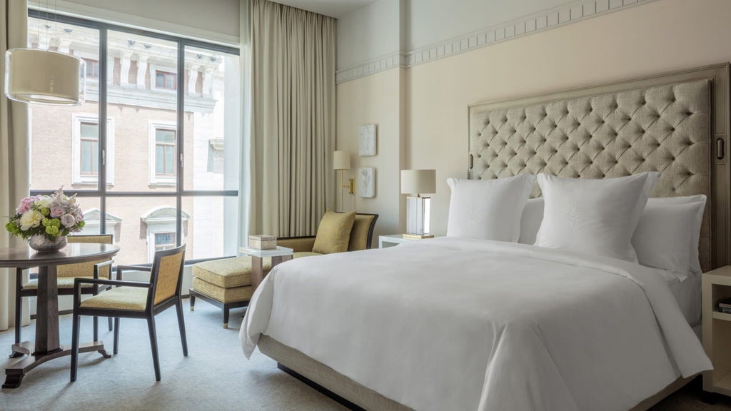 Spacious luxury hotel room with gold accents featuring king bed, ornate headboard, crystal chandelier and floor-to-ceiling windows with city views