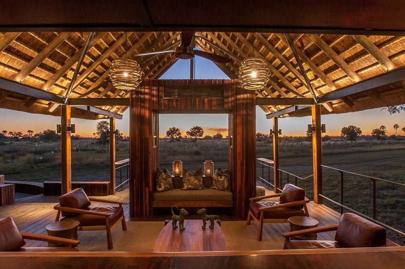 Elevated safari lodge with thatched-roof cottages overlooking a vast savanna, connected by wooden walkways amidst acacia trees