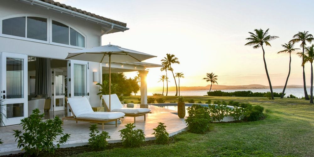 Luxurious Estate Suite at Jumby Bay, featuring elegant four-poster bed, white linens, and expansive ocean view through large windows in Antigua's tropical paradise.