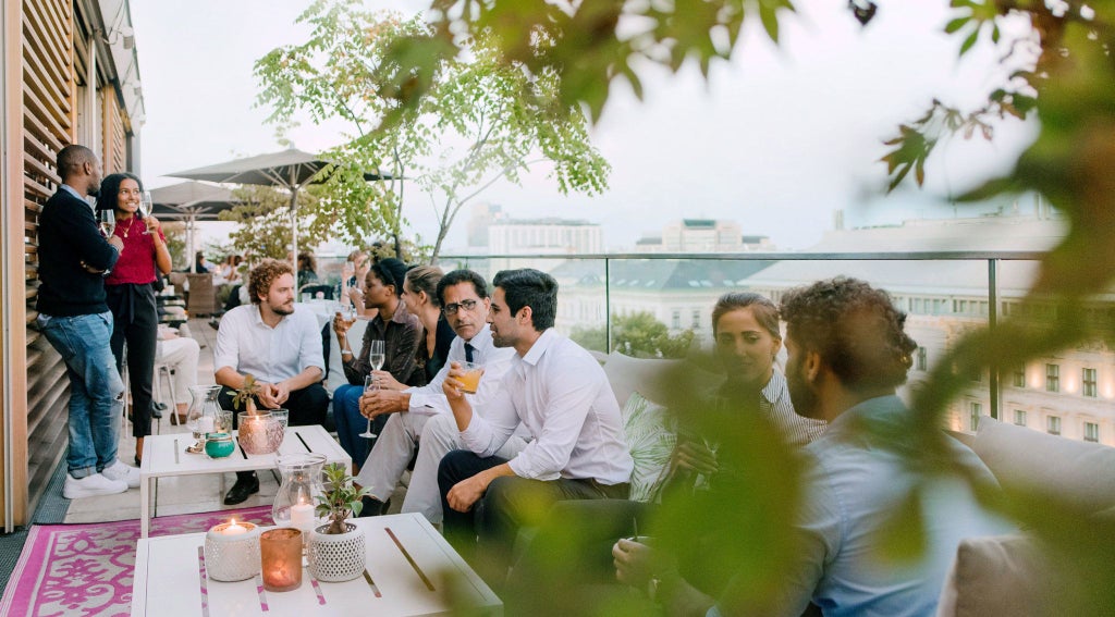 Elegant rooftop terrace of Grand Ferdinand hotel overlooking Vienna skyline, with plush white lounge chairs and panoramic alpine mountain views at sunset