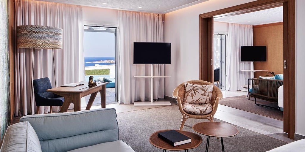 Luxurious Mediterranean suite with white-washed walls, blue accents, private balcony overlooking Aegean Sea, elegant modern decor, and sweeping Mykonos landscape view