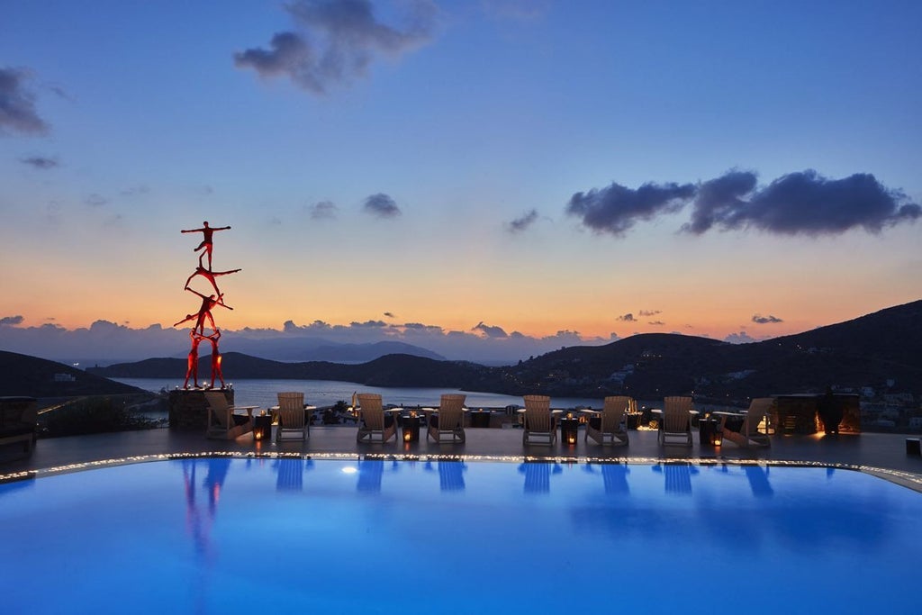 Luxurious white hotel complex with infinity pool overlooking Aegean Sea at sunset, surrounded by traditional Greek architecture and palm trees.