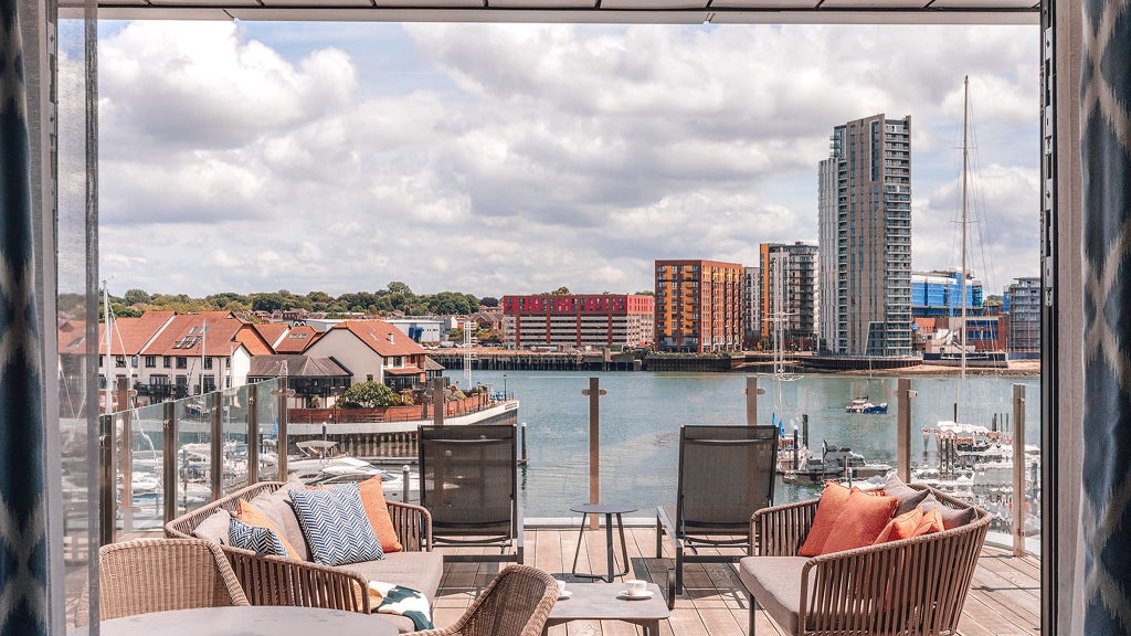 Modern maritime-style suite with expansive glass doors opening to a balcony overlooking Southampton's marina, featuring elegant contemporary furnishings and soft neutral tones