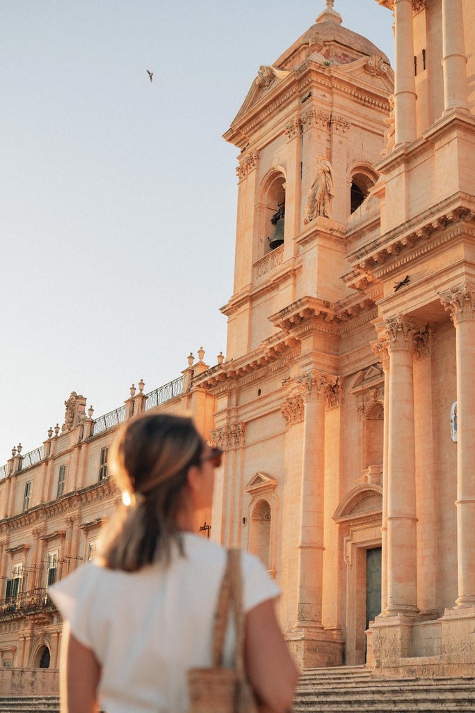 Elegant Sicilian boutique hotel with ornate balconies, warm terracotta facade, and lush green potted plants against historic stone architecture