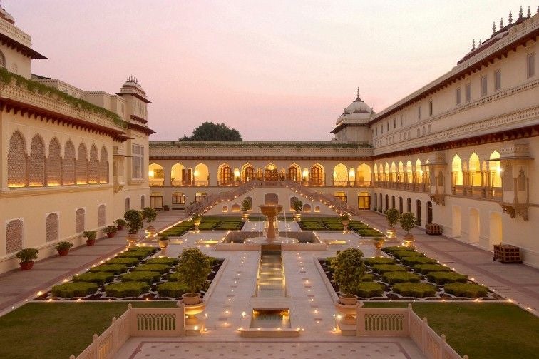 Ornate sandstone Indian palace hotel with grand domed architecture, manicured gardens, and reflection pool under a golden sunset sky.